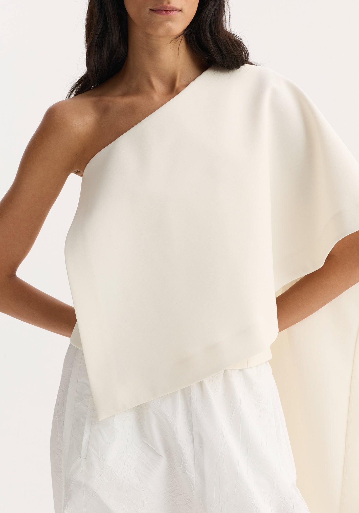 Model wearing Draped Cape Top | Cream with a single sleeve and elegant cape-like panel, paired with loose textured white bottoms, highlighting its refined silhouette.