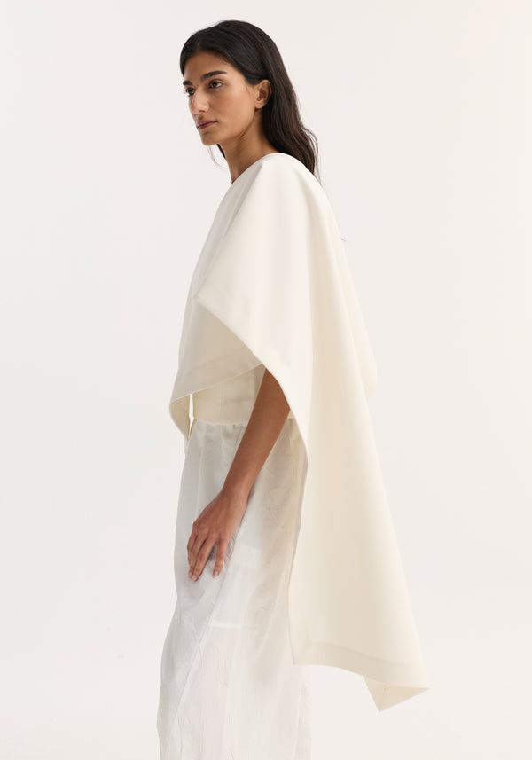 Model in profile wearing Draped cape top | cream with single sleeve and flowing scarf detail, paired with a long skirt, creating an elegant, minimalist look.