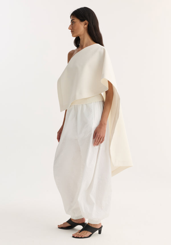 Model in profile wearing Draped cape top | cream, featuring a single sleeve and long draped scarf detail, paired with high-waisted balloon trousers.