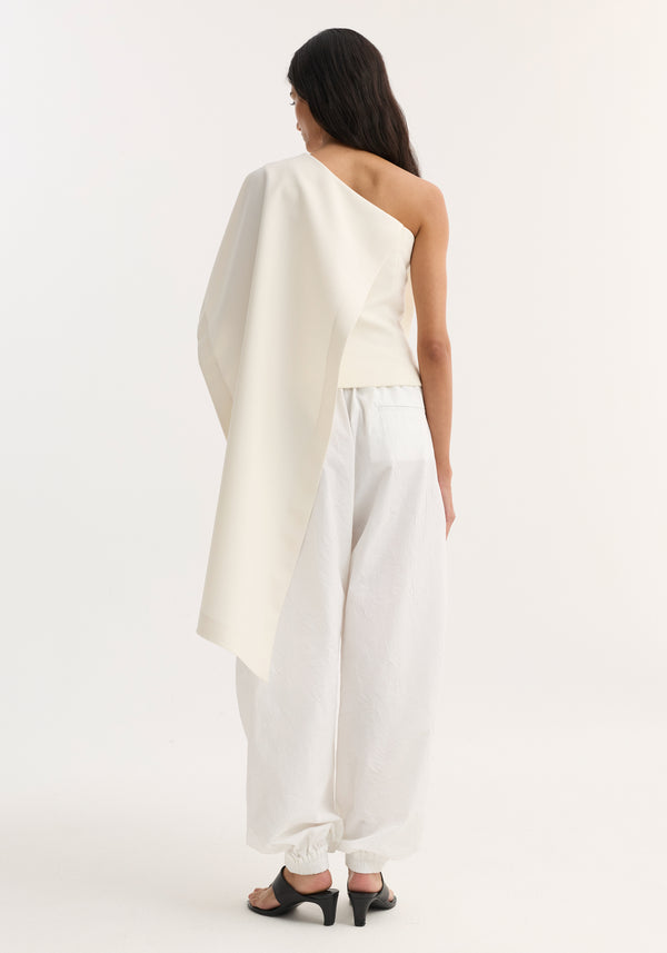 Woman in a Draped Cape Top | Cream, featuring an asymmetrical single sleeve and long draped panel, paired with textured trousers and heeled sandals.