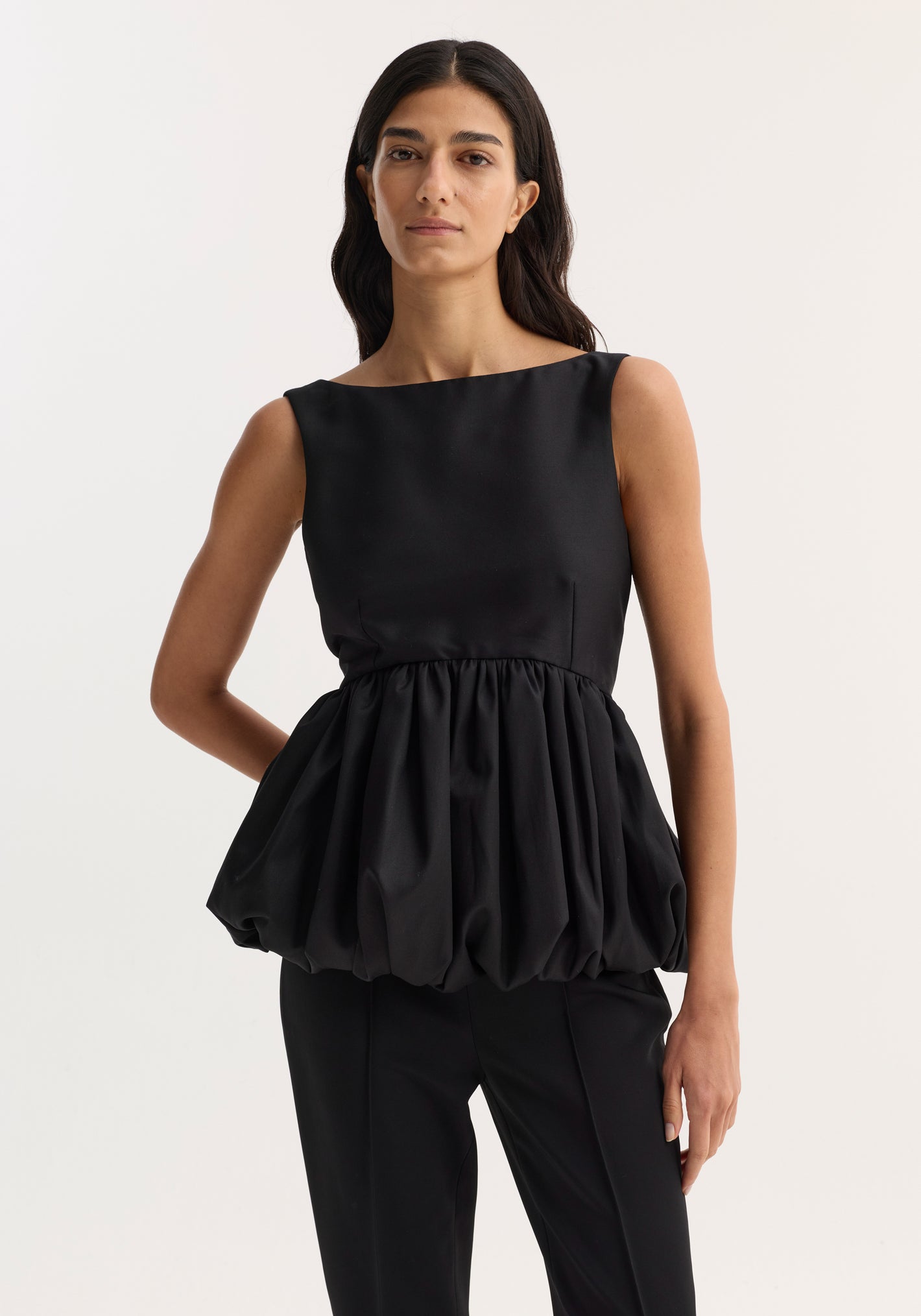 Voluminous boat-neck top in black, featuring a sleeveless design with a low back and peplum waist, worn by a slim, dark-haired woman.
