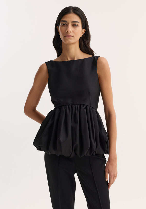 Voluminous boat-neck top in black, featuring a sleeveless design with a low back and peplum waist, worn by a slim, dark-haired woman.