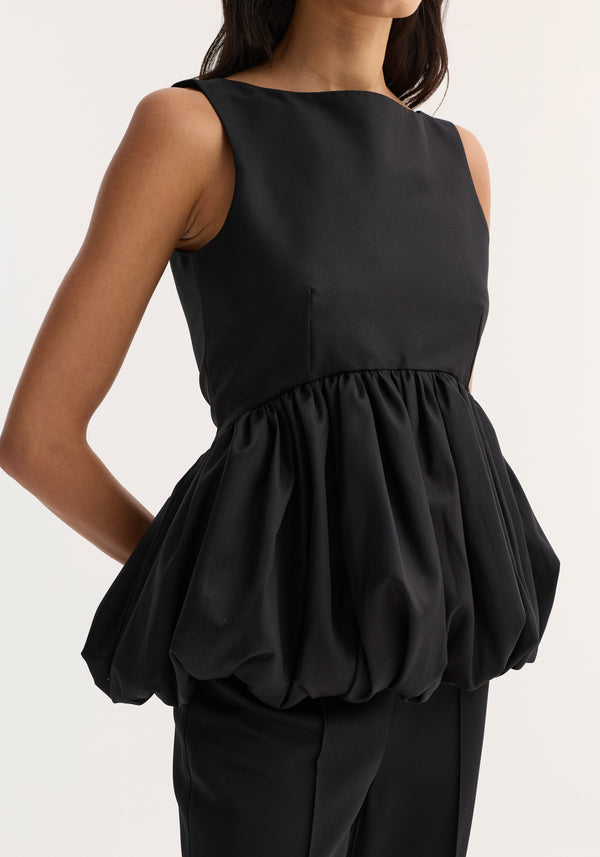 Woman in a Voluminous boat-neck top | black, featuring a sleeveless design, high neckline, and a dramatic peplum over slim trousers, creating a sleek silhouette.