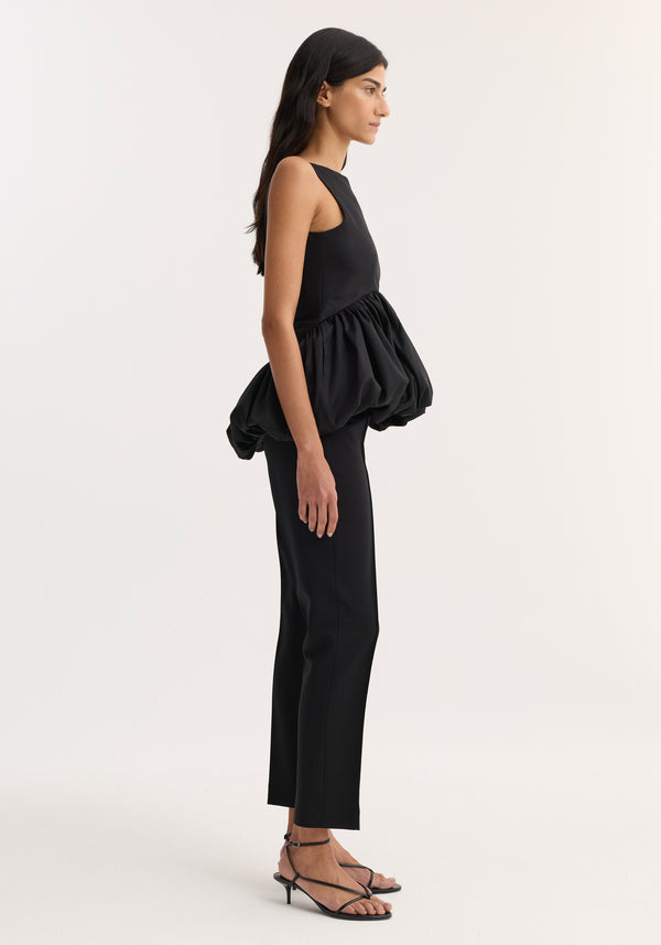 Model wearing a Voluminous boat-neck top in black with a sculptural peplum, paired with straight-leg trousers and strappy kitten-heel sandals.