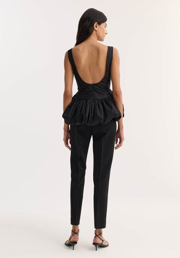 Voluminous boat-neck top in black, featuring a sleeveless design with a low scooped back and a sculptural peplum hem, worn with tailored trousers.