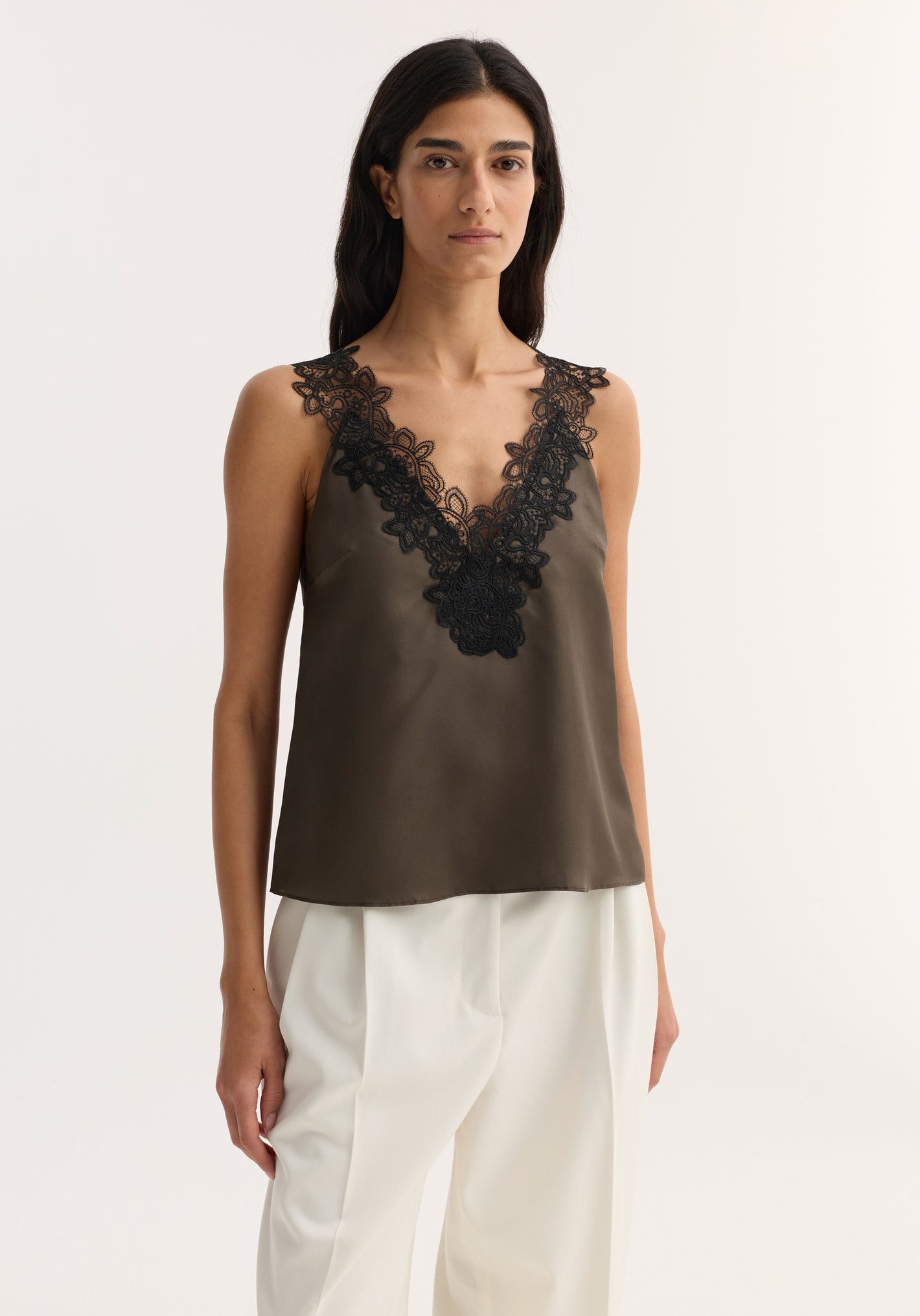 Woman in V-neck silk lace top | earth, featuring a sleeveless design with intricate lace-trimmed V-neck, paired with high-waisted pleated trousers.
