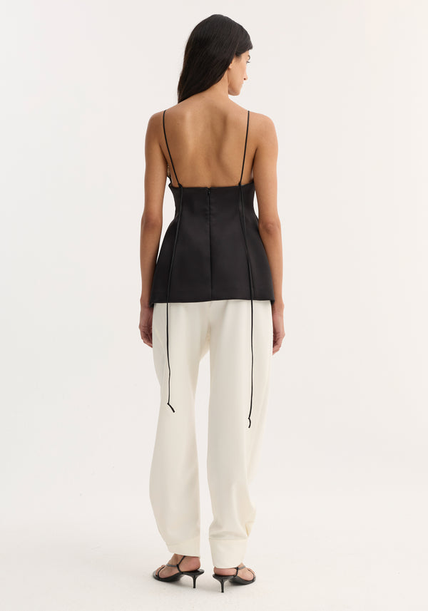 Woman in a Bustier top | black, featuring thin straps and bustier stitching, paired with high-waisted white trousers. Straight dark hair enhances the minimalist style.