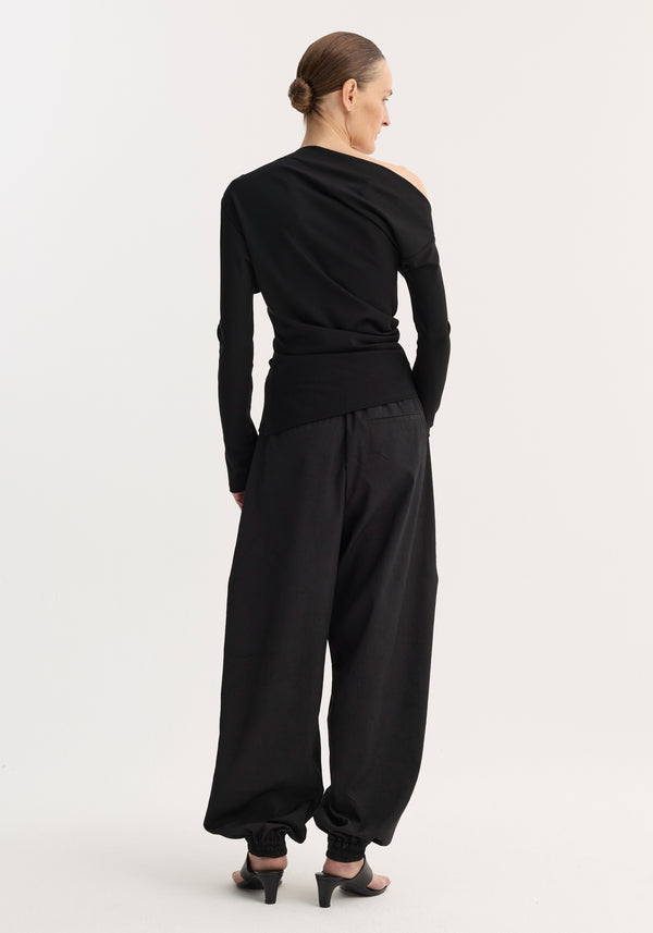 Woman in a fitted black Twisted Jersey Top with one shoulder exposed, paired with loose black trousers and open-toe mules, hair in a bun.