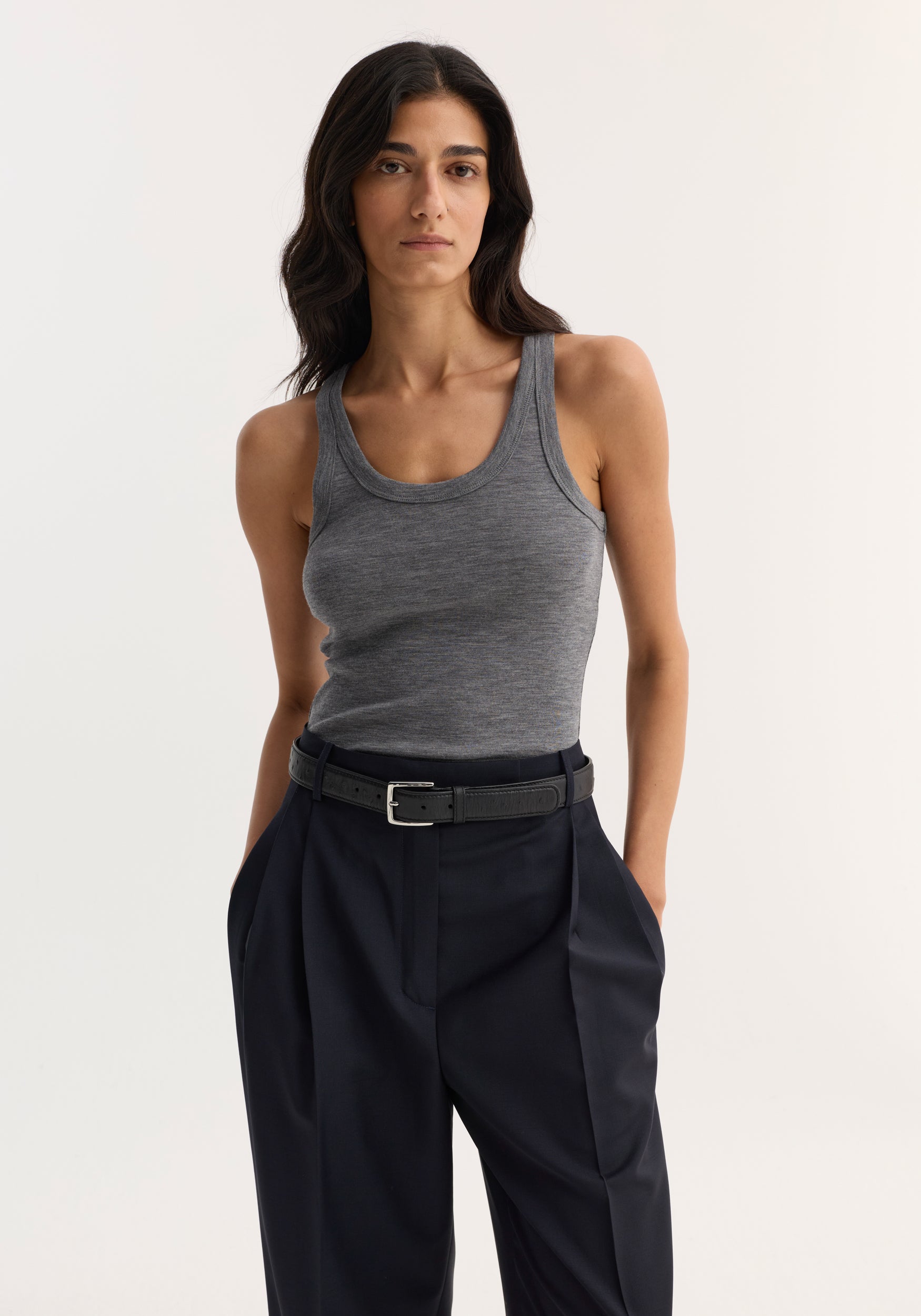 Model wearing Merino scoop neck top in mid grey melange, tucked into high-waisted trousers, showcasing its sleeveless, fitted design crafted from 100% merino wool.