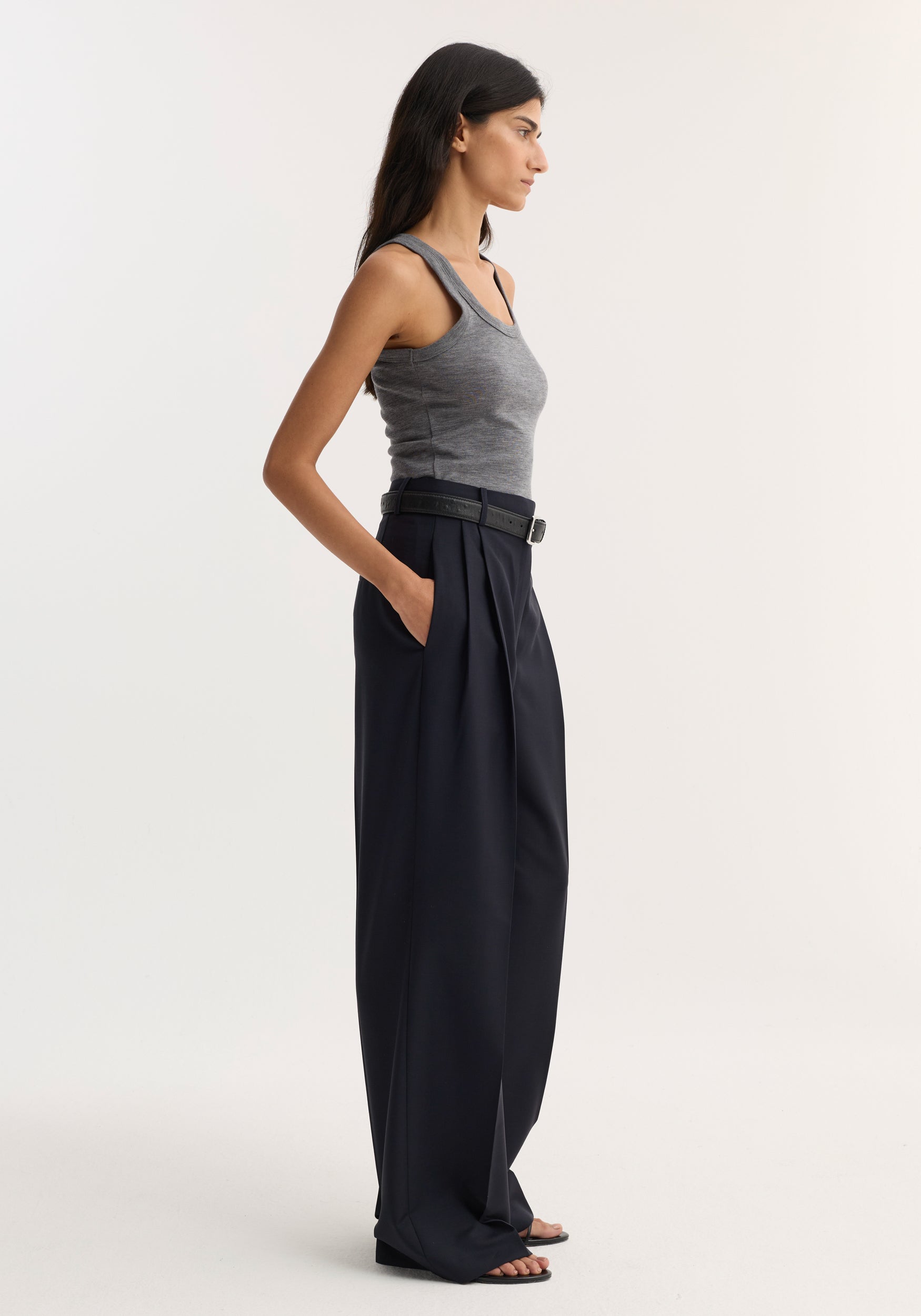 Model in profile wearing a Merino scoop neck top, mid grey melange, tucked into high-waisted black trousers, accessorized with a black belt and sandals.