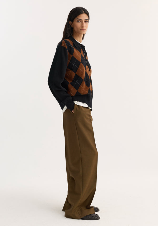 Model wearing a Compact knitted check polo in black rust check, featuring an argyle pattern, structured collar, and three-button front, paired with high-waisted trousers.