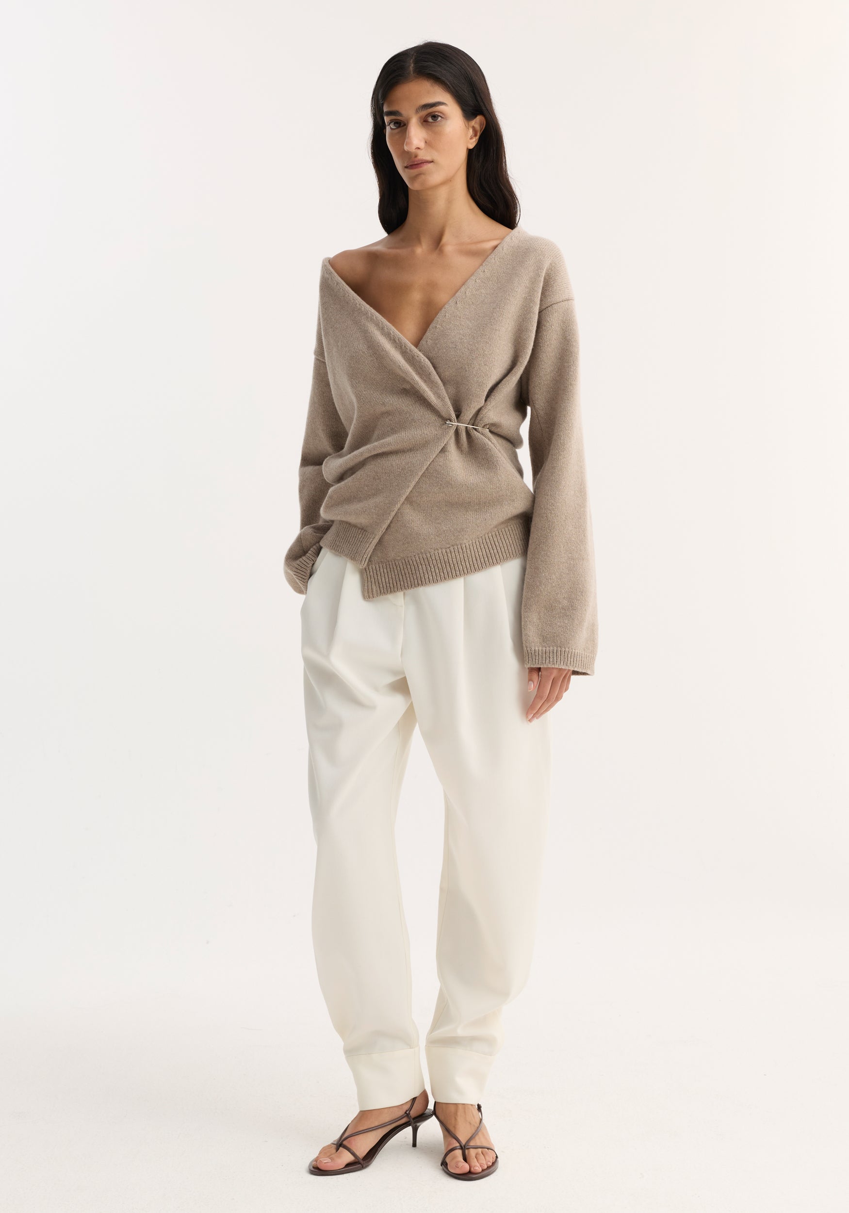 Model in Off-shoulder wrap cardigan | light taupe, wool-cashmere blend, with safety pin closure, paired with high-waisted white trousers and brown strappy sandals.