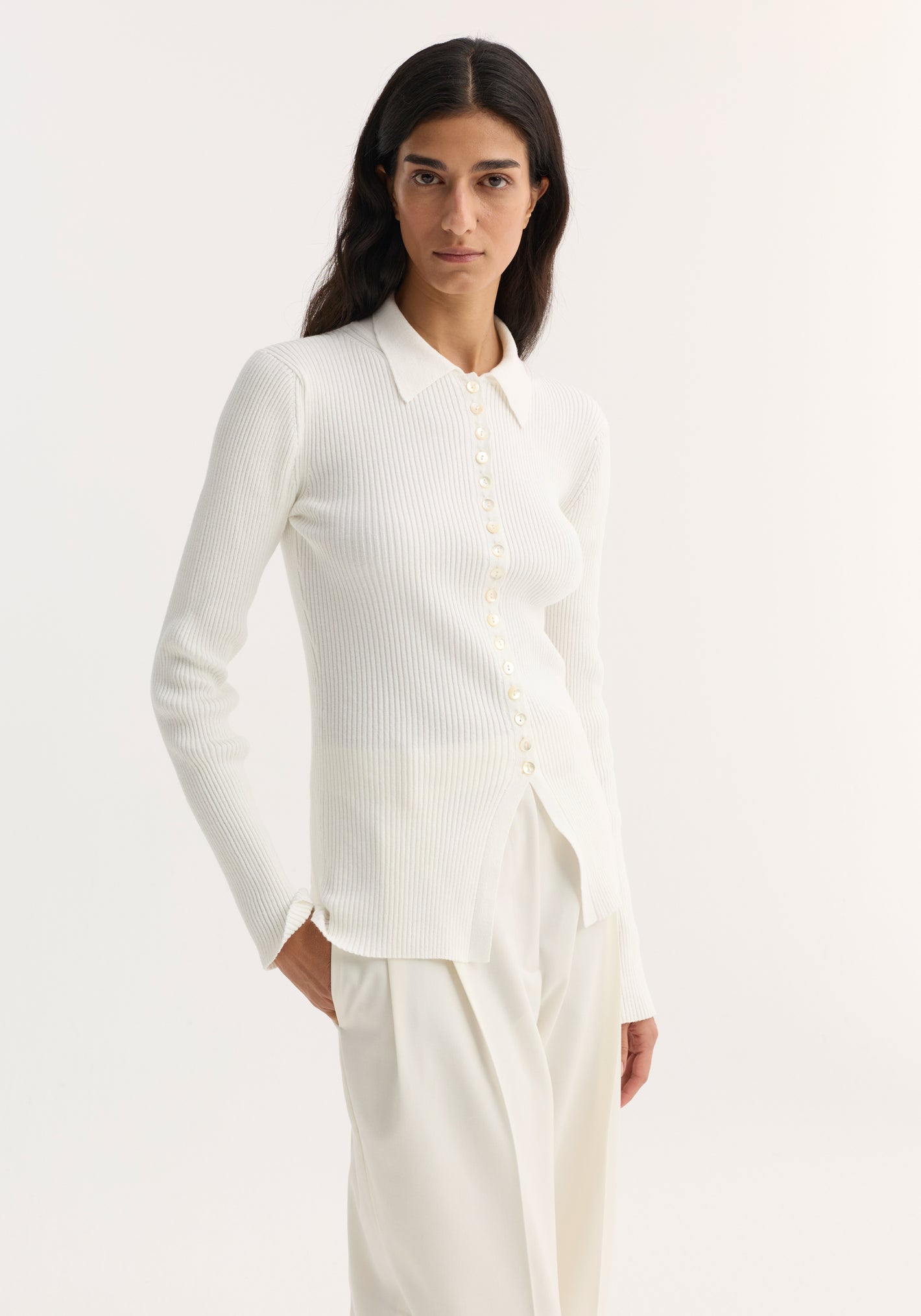 Woman wearing Elongated rib cardigan in off-white, featuring a pointed collar, button closures, and a ribbed texture, paired with high-waisted trousers.