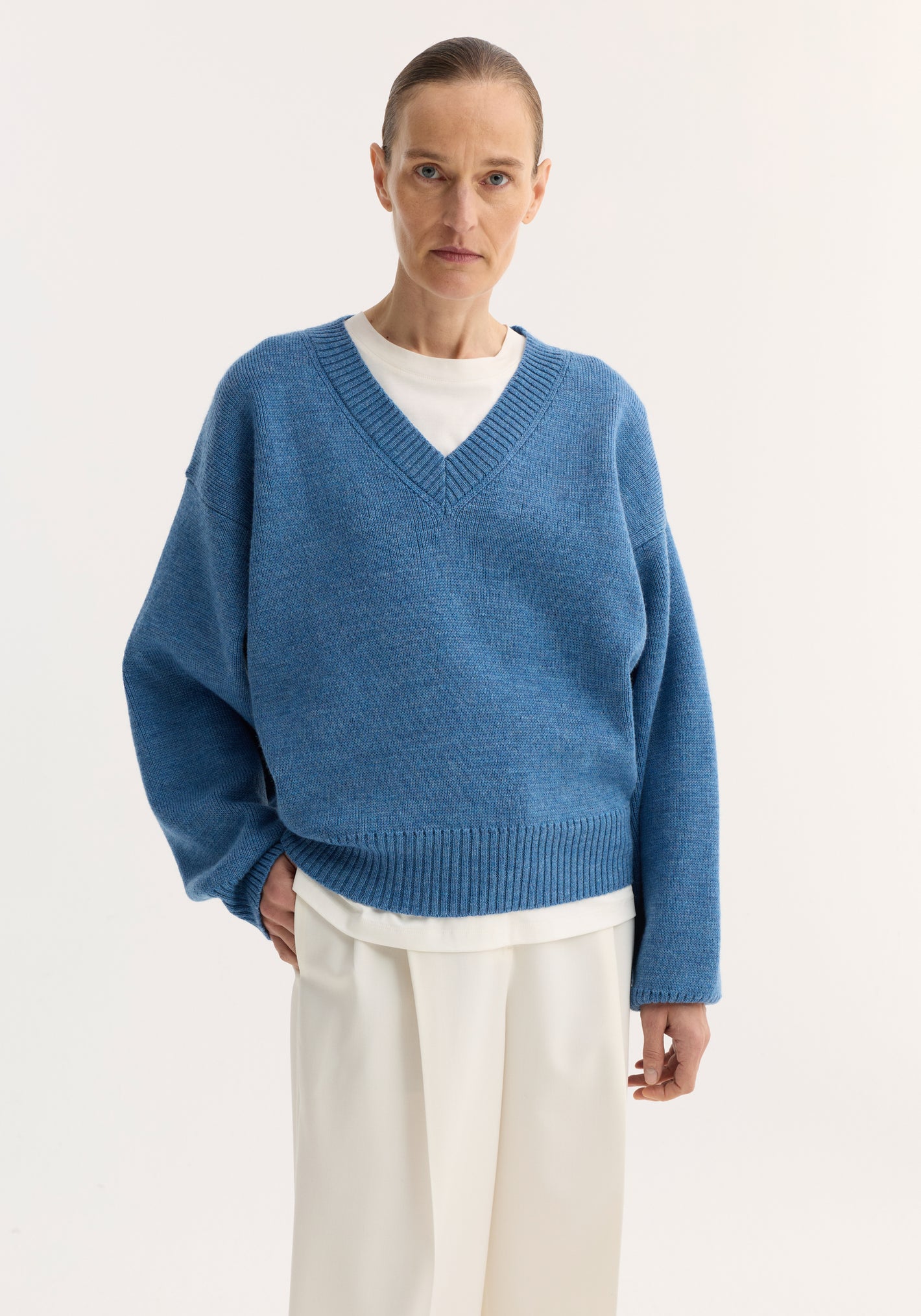 Person wearing a V-neck knitted jumper in denim blue melange, layered over a white shirt, paired with cream wide-leg trousers. Relaxed fit and dropped shoulders.