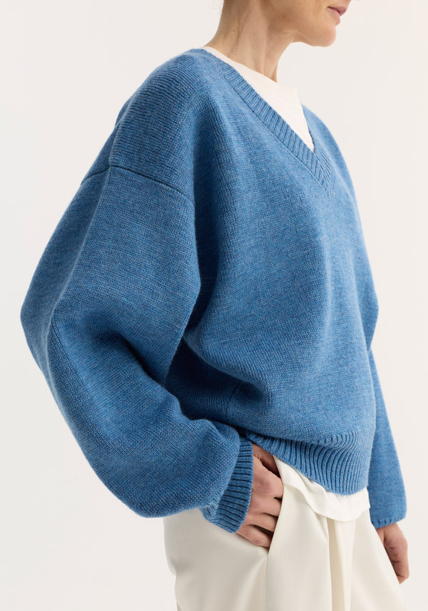 V-neck knitted jumper in denim blue melange, featuring a relaxed fit with dropped shoulders, layered over a white top, paired with off-white trousers.