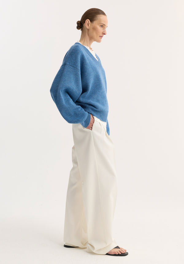 Side profile of a person wearing a denim blue melange V-neck knitted jumper with a relaxed fit, paired with cream wide-leg trousers and black sandals.