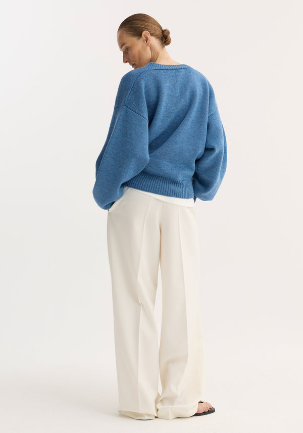 Person in V-neck knitted jumper, denim blue melange, with relaxed fit and dropped shoulders, paired with high-waisted white trousers and black sandals, hair in bun.