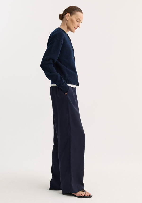 Model in profile wearing Short boiled cardigan, navy, with high crew neck and drop shoulders, over a white top, paired with matching navy trousers and black sandals.
