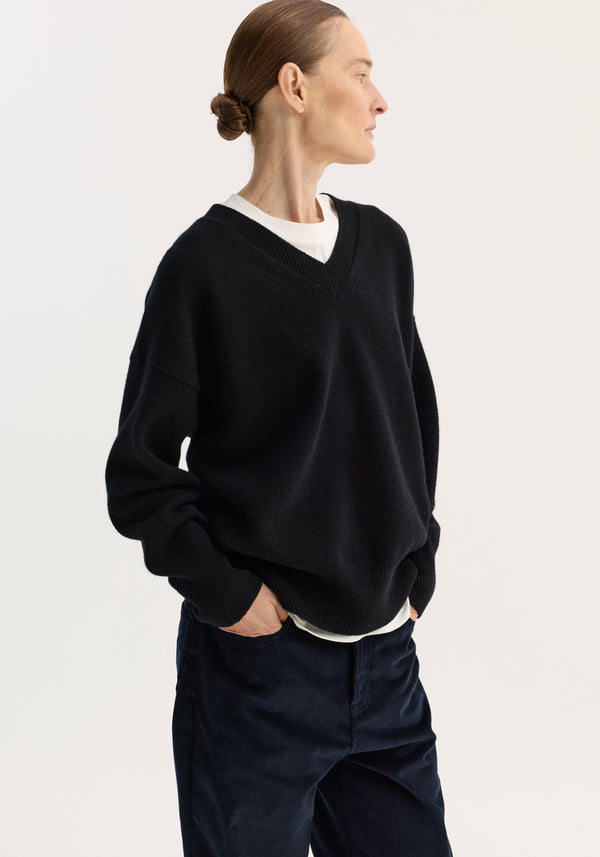 Person wearing a V-neck knitted jumper in black, featuring a relaxed fit and drop shoulders, over a white T-shirt, paired with dark corduroy pants.