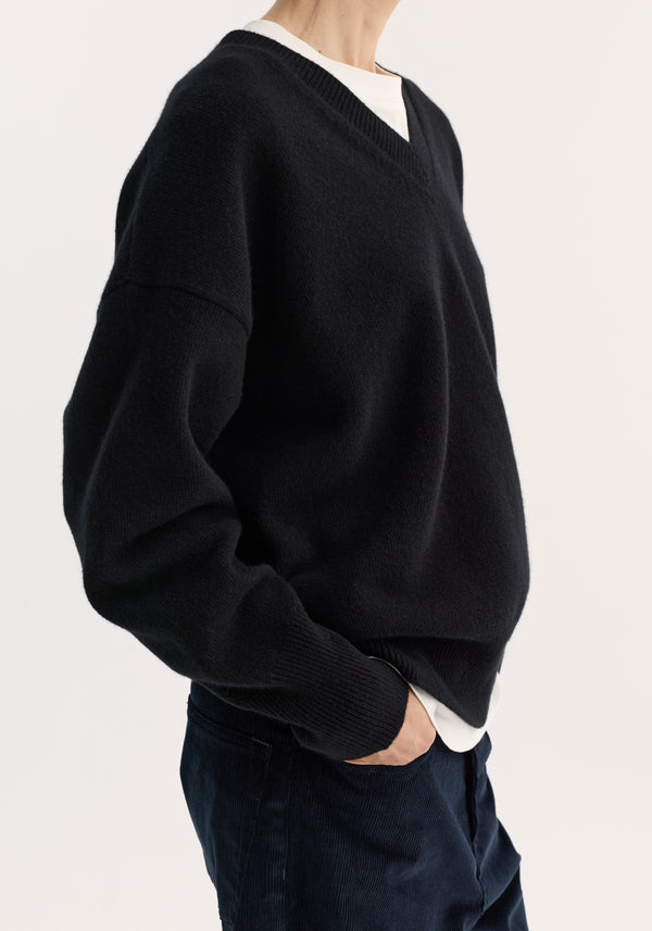 Person wearing a black V-neck knitted jumper over a white T-shirt, showcasing its relaxed fit and dropped shoulders, with one hand in pocket of dark trousers.
