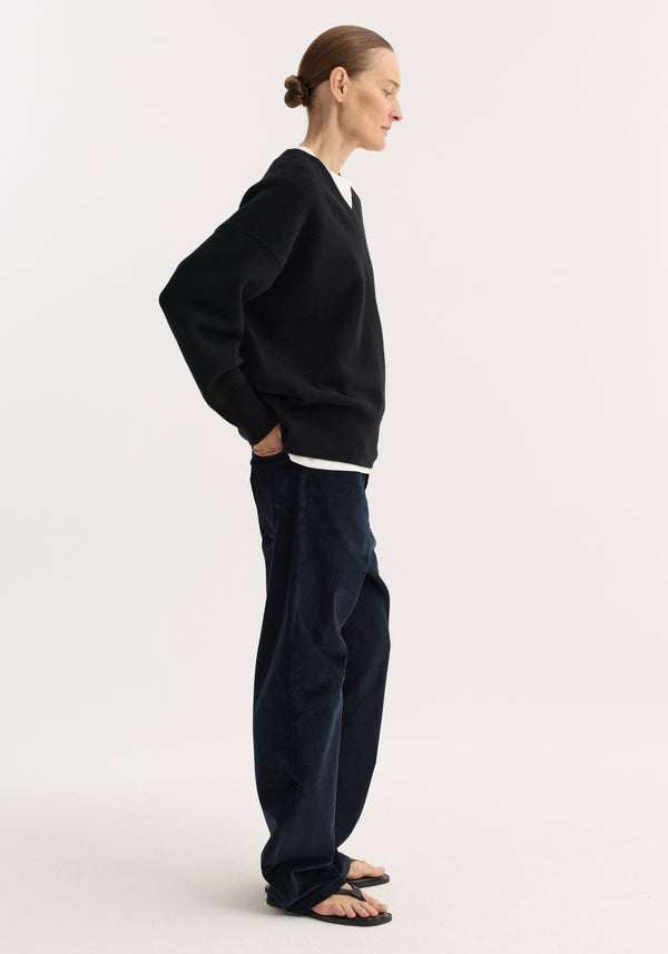 V-neck knitted jumper in black, oversized with a relaxed fit and dropped shoulders, worn by model with hands in pockets, showcasing minimalist style.