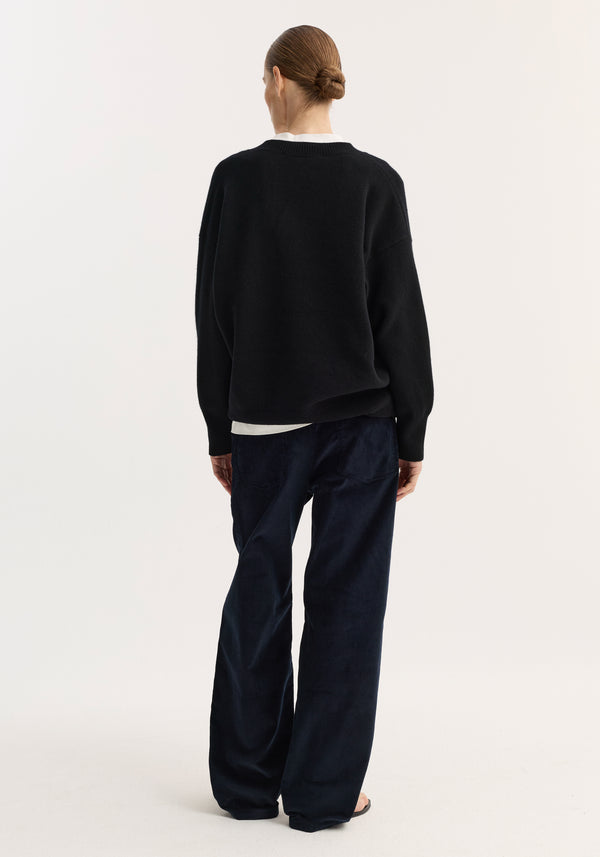 Person wearing a loose-fitting V-neck knitted jumper in black, with a relaxed fit and dropped shoulders, paired with wide-leg trousers. Hair styled in a low bun.