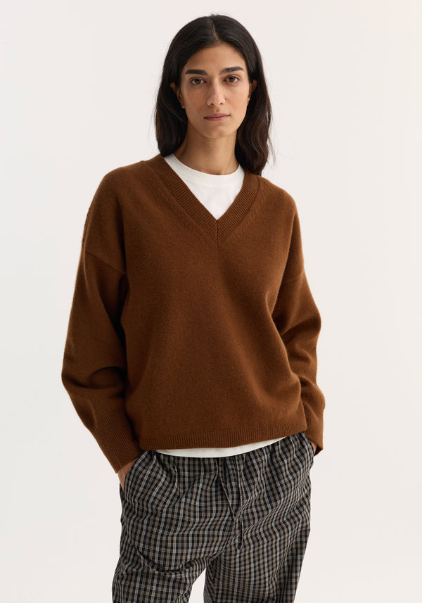 Young woman models a V-neck knitted jumper in rust, showcasing its relaxed fit, drop shoulders, and soft wool blend, paired with plaid trousers.