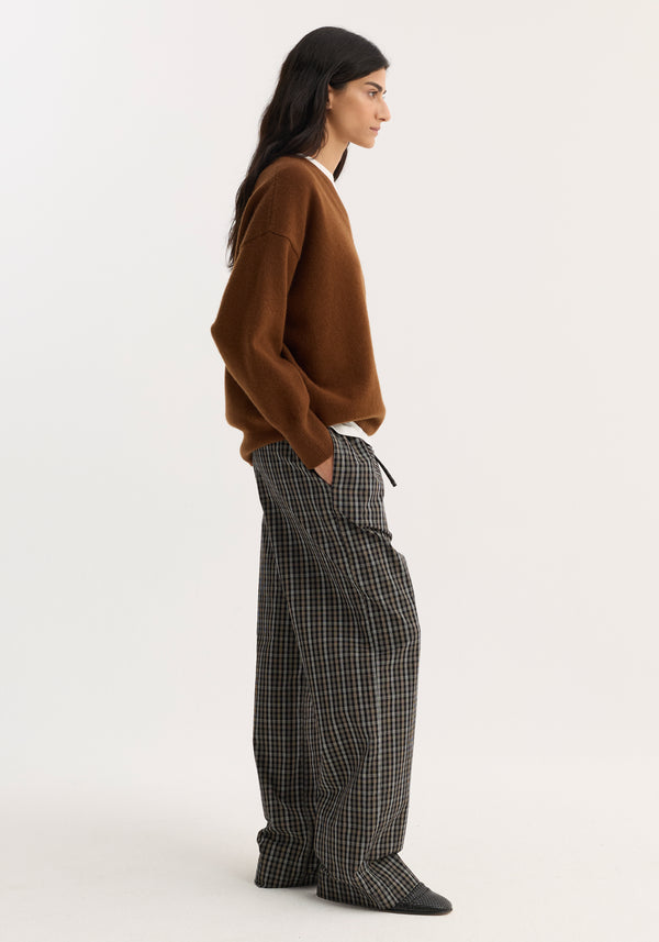 Model in V-neck knitted jumper, rust, with relaxed fit, dropped shoulders, layered over a white T-shirt, paired with plaid trousers and black flats.