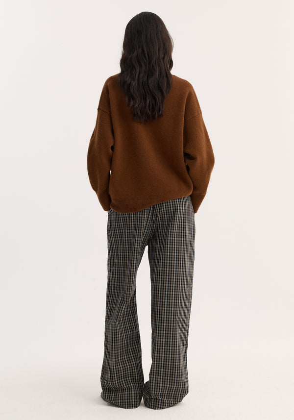 Person with long hair wearing a V-neck knitted jumper in rust, featuring a relaxed fit and dropped shoulders, paired with wide-leg plaid trousers.