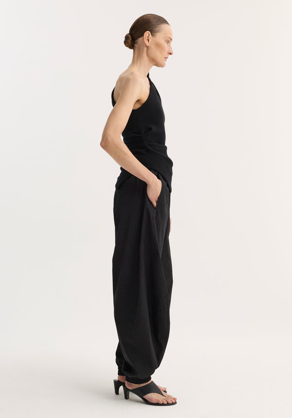Model wearing Off-shoulder top | black, featuring a one-shoulder design, fitted waist, and asymmetrical hem, paired with high-waisted trousers and heeled sandals.