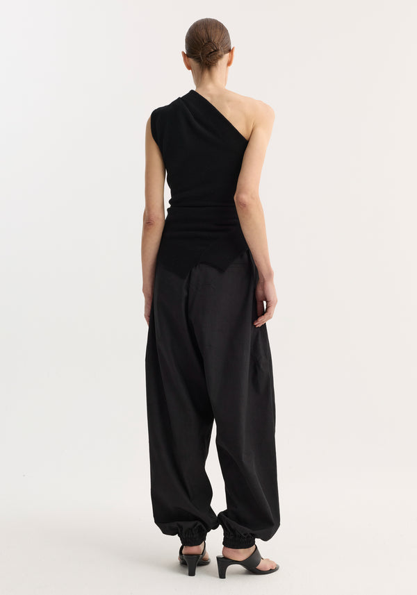 Model in Off-shoulder top | black, featuring a fitted, one-shoulder design with asymmetrical hem, paired with high-waisted trousers and open-toe mules, hair in low bun.