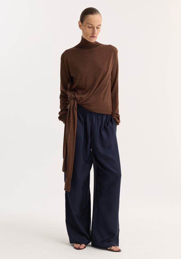 Model in a 4-sleeved knitted jumper | brown, featuring a turtleneck and tie sleeve detail, paired with navy wide-leg trousers and sandals.