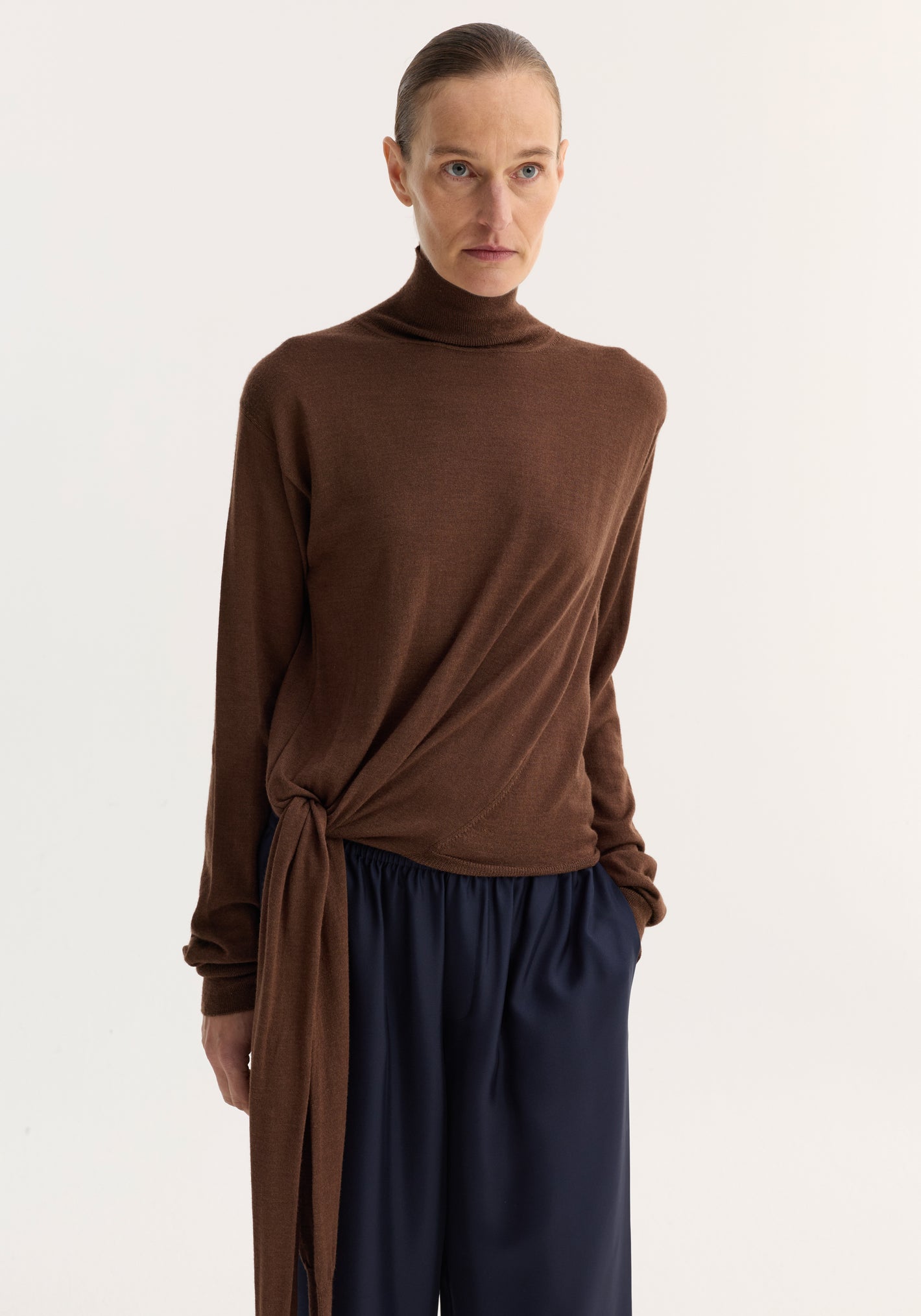 Light-skinned model wears 4-sleeved knitted jumper | brown, showcasing a turtleneck with tie sleeve detail, paired with loose navy trousers in a studio setting.