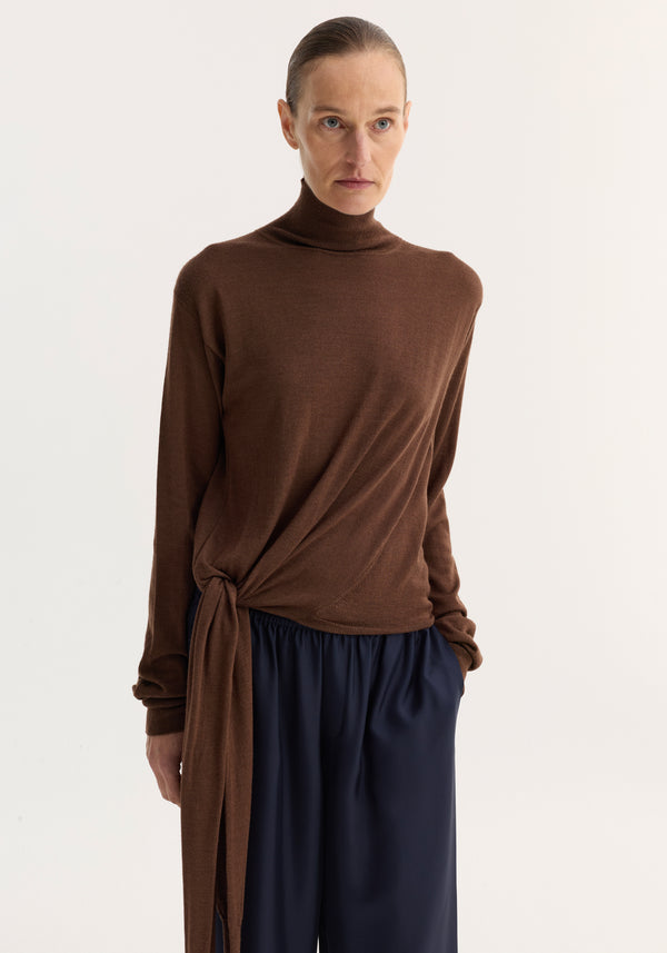 Light-skinned model wears 4-sleeved knitted jumper | brown, showcasing a turtleneck with tie sleeve detail, paired with loose navy trousers in a studio setting.