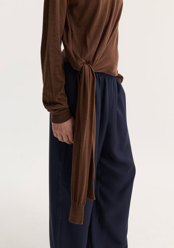 Model wearing 4-sleeved knitted jumper | brown, showcasing its knotted hip design and sculptural tie sleeve detail over high-waisted navy trousers.