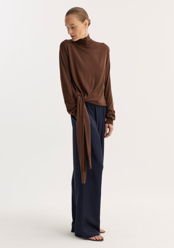 Model in profile wearing the 4-sleeved knitted jumper | brown, featuring a gathered sleeve layer tied at the hem, paired with wide-leg navy trousers.