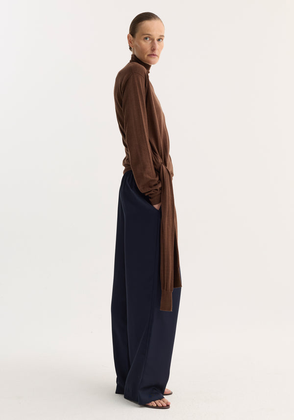 Model wearing a 4-sleeved knitted jumper | brown, featuring a unique tie sleeve detail, paired with wide-leg trousers and flat sandals.