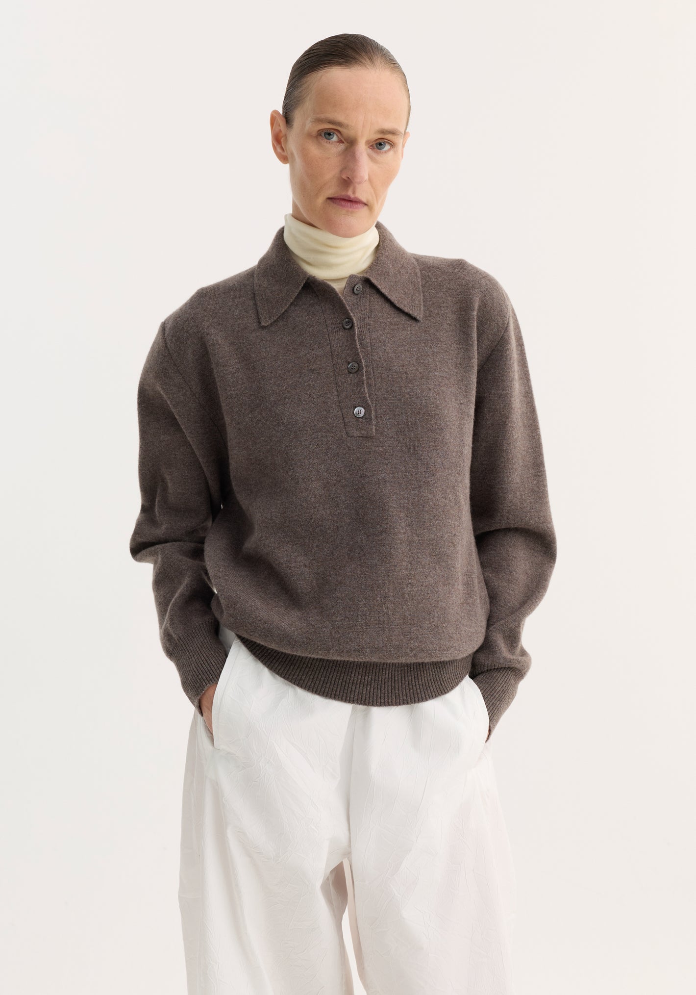 Person wearing Compact knitted polo | mud melange, featuring a structured collar and three-button placket, layered over a turtleneck with white trousers.
