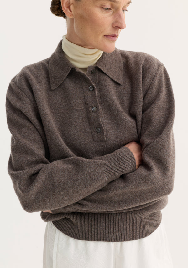 Person wearing Compact Knitted Polo in mud melange with a structured collar and three-button closure over a cream turtleneck, arms folded.