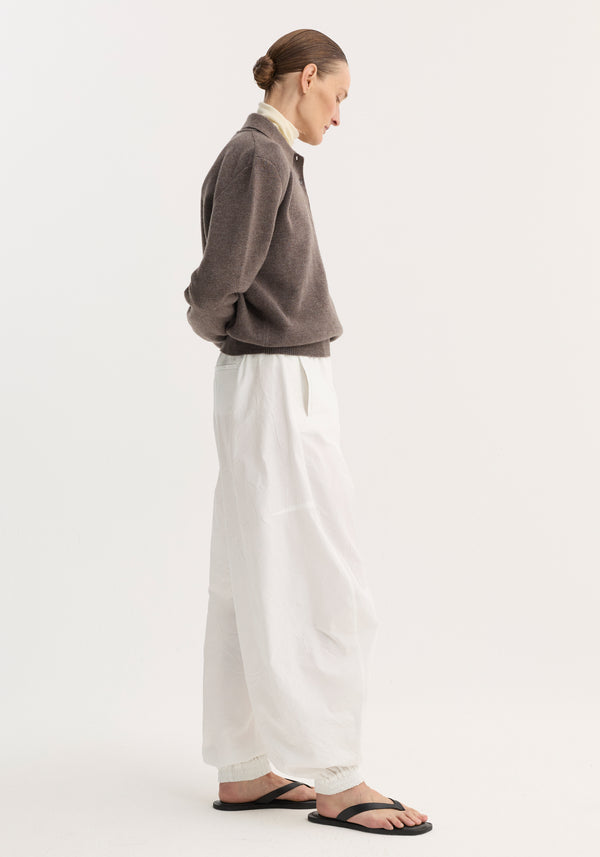 Side-profile of a model in a Compact knitted polo, mud melange, featuring a high neck and relaxed fit, paired with wide, billowy trousers.