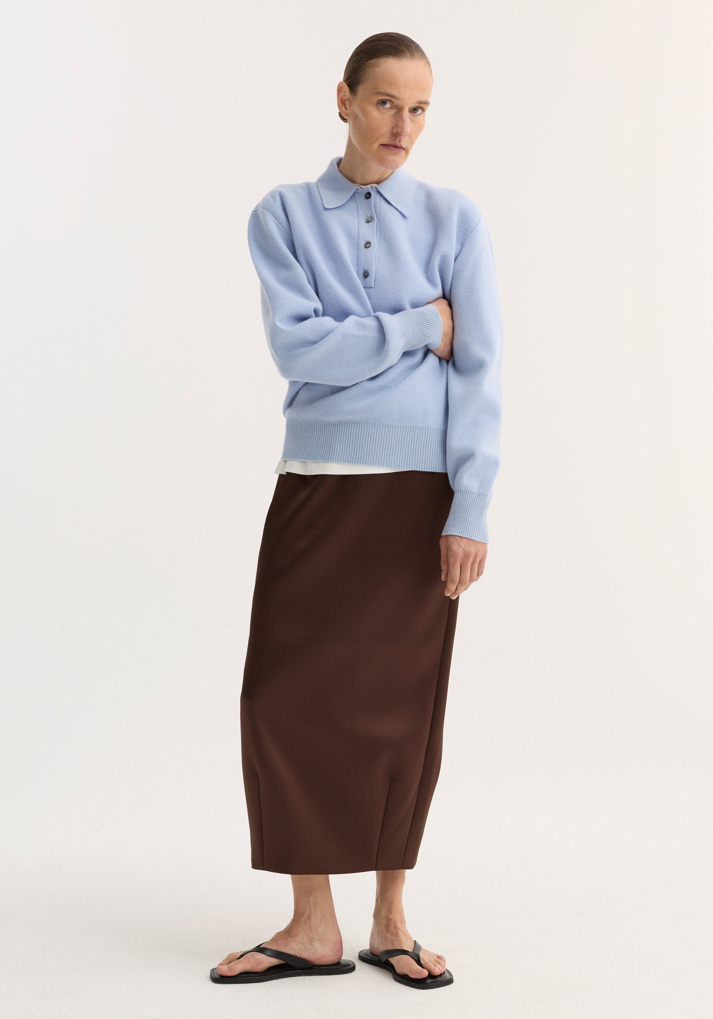 Model in baby blue compact knitted polo with collar and button placket, layered over white, paired with deep brown ankle-length skirt and black flat sandals.