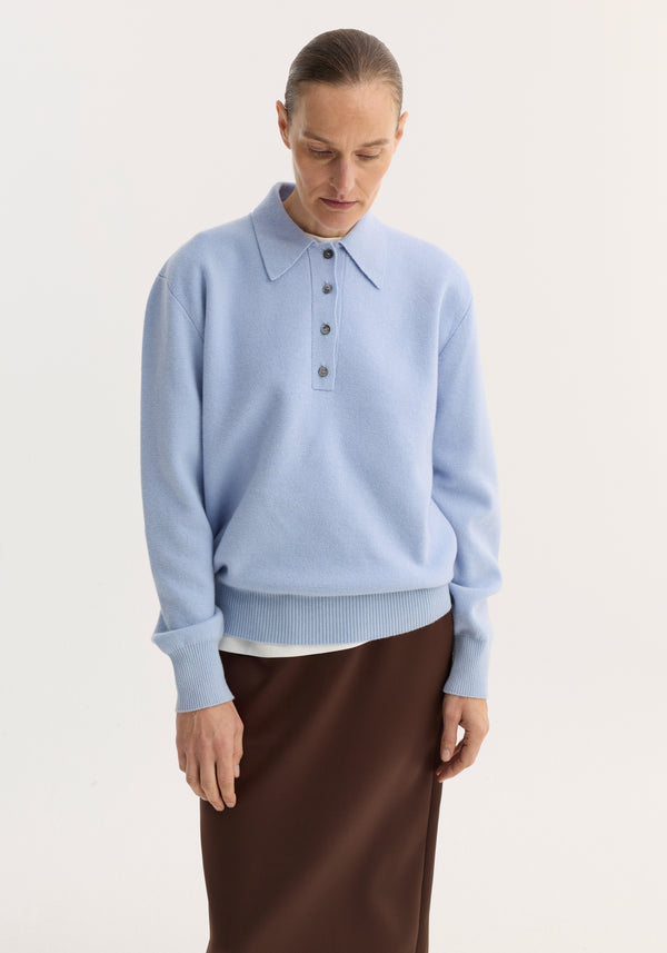 Person wearing a baby blue compact knitted polo with a pointed collar and four-button placket, paired with a brown skirt, looking downward.