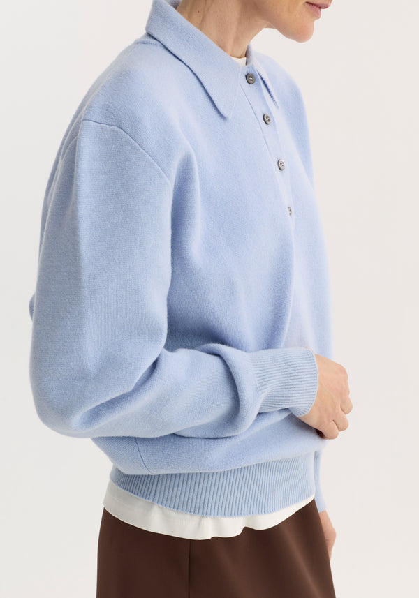 Model wearing a Compact knitted polo in baby blue, featuring a long-sleeved design with a polo collar and 3-button front, layered over a white top.
