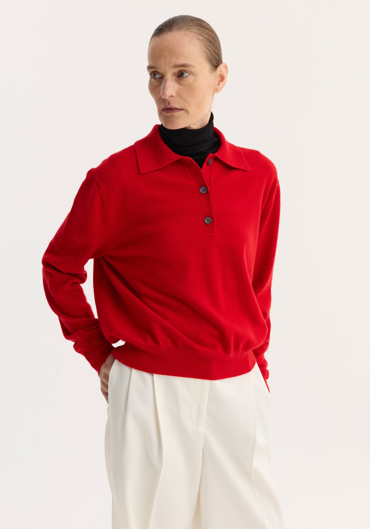 Model in a Wool Cashmere Polo | Red, featuring a relaxed fit, structured collar, and three-button closure, layered over a black turtleneck with white trousers.