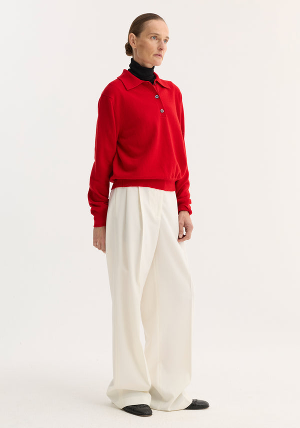 Model wears Wool Cashmere Polo | Red over a black turtleneck, paired with high-waisted white trousers and black loafers, highlighting the sweater's structured collar and buttons.