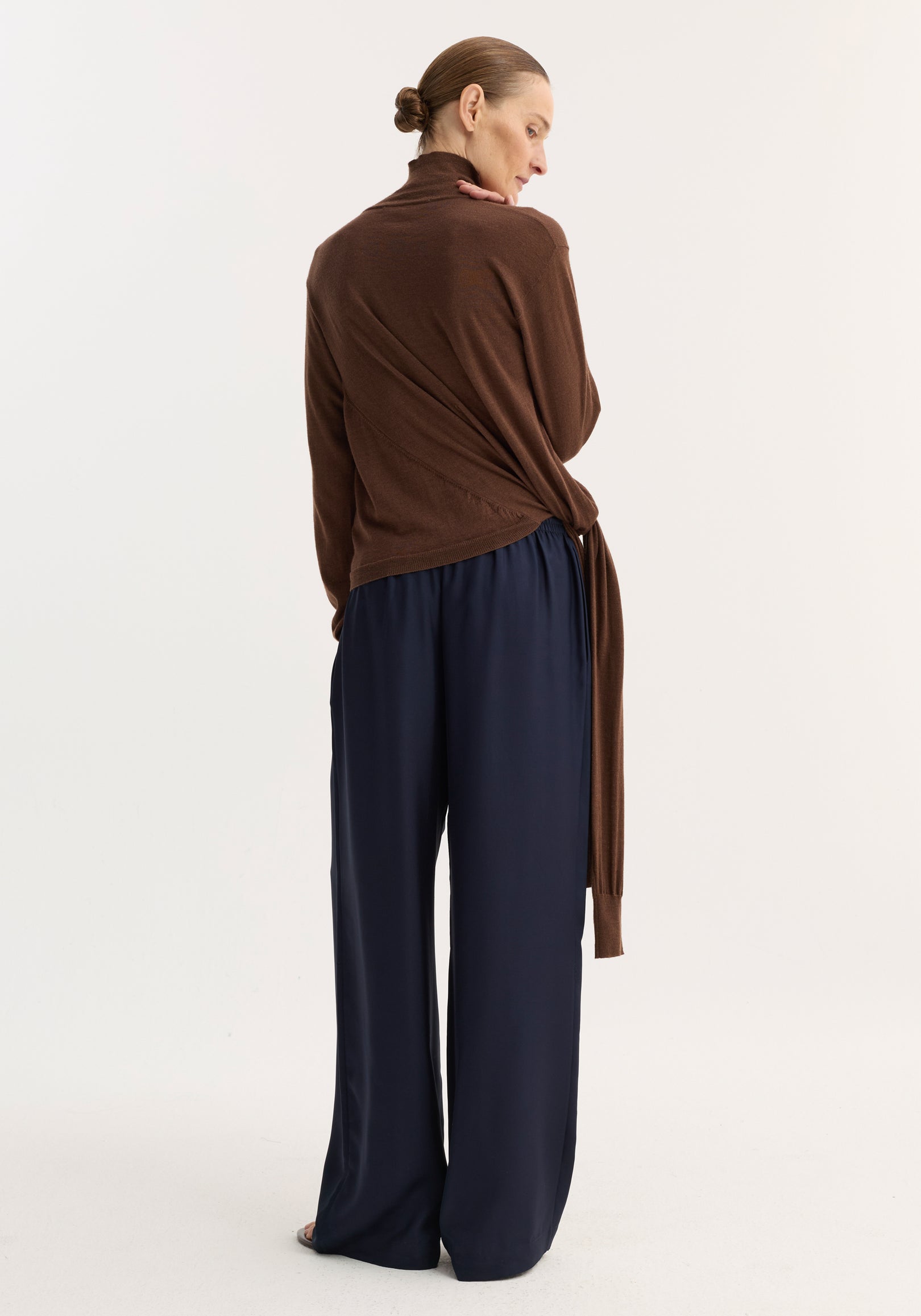 Woman wearing wide leg silk trousers with elasticated waistband and drawstring, styled with a brown turtleneck sweater, showcasing the trousers' relaxed drape and fluidity.