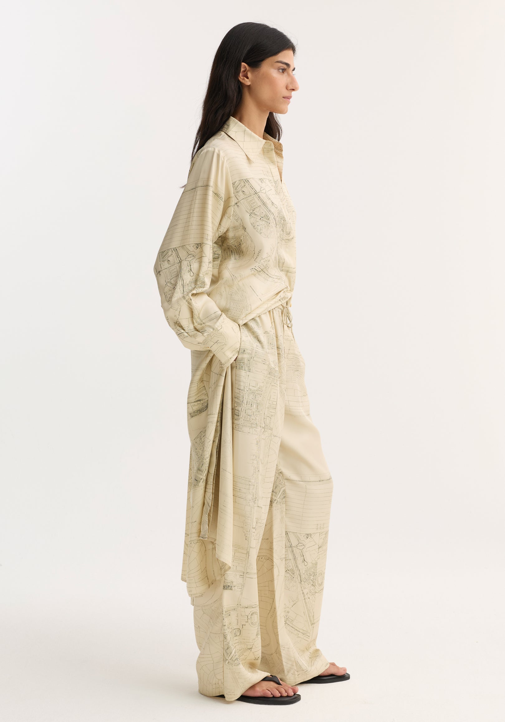 Person in profile wearing relaxed, oversized wide leg silk trousers with Amsterdam map print, featuring an elasticated waistband and drawstring. Paired with black flip-flops.