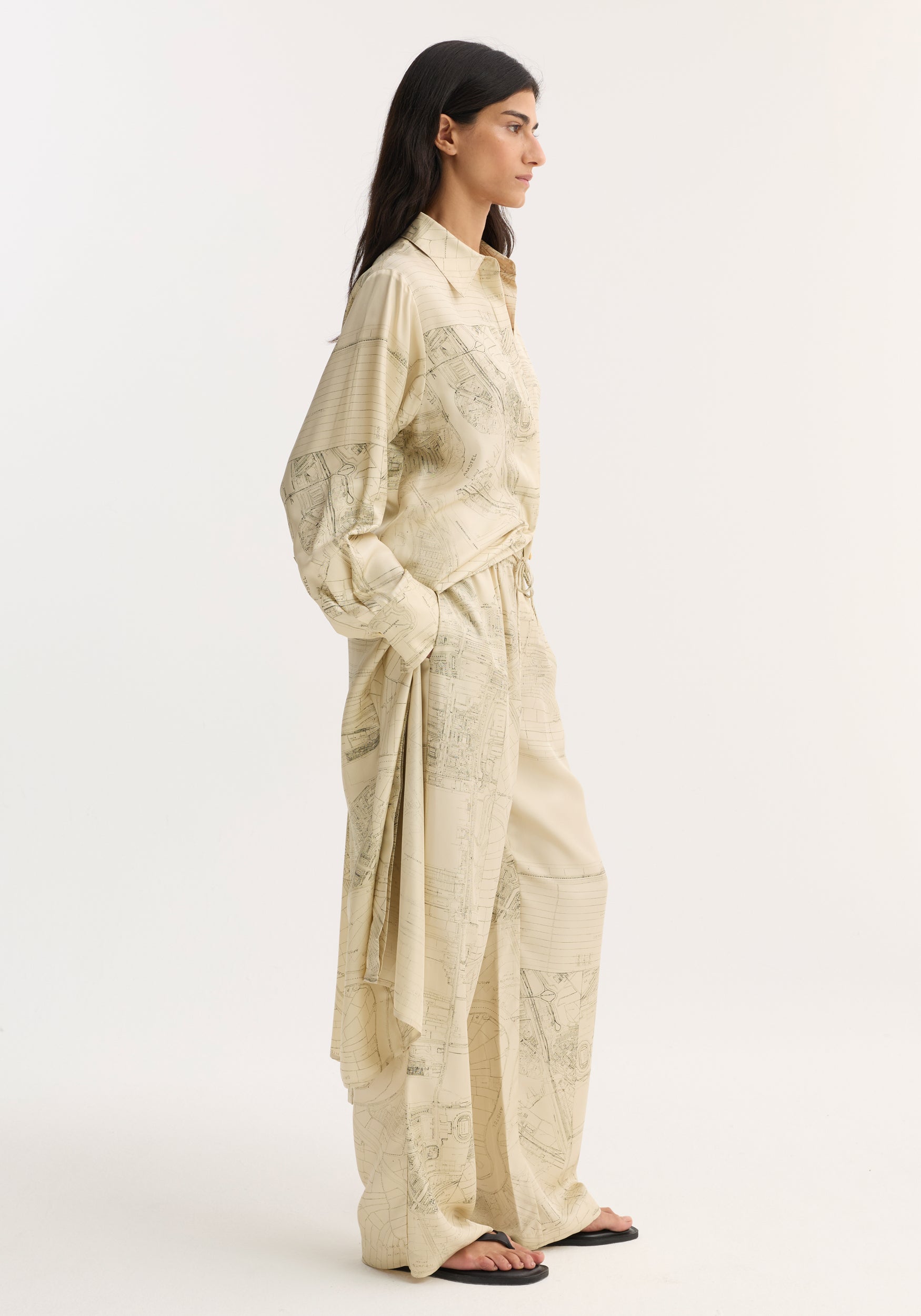 Person in profile wearing relaxed, oversized wide leg silk trousers with Amsterdam map print, featuring an elasticated waistband and drawstring. Paired with black flip-flops.