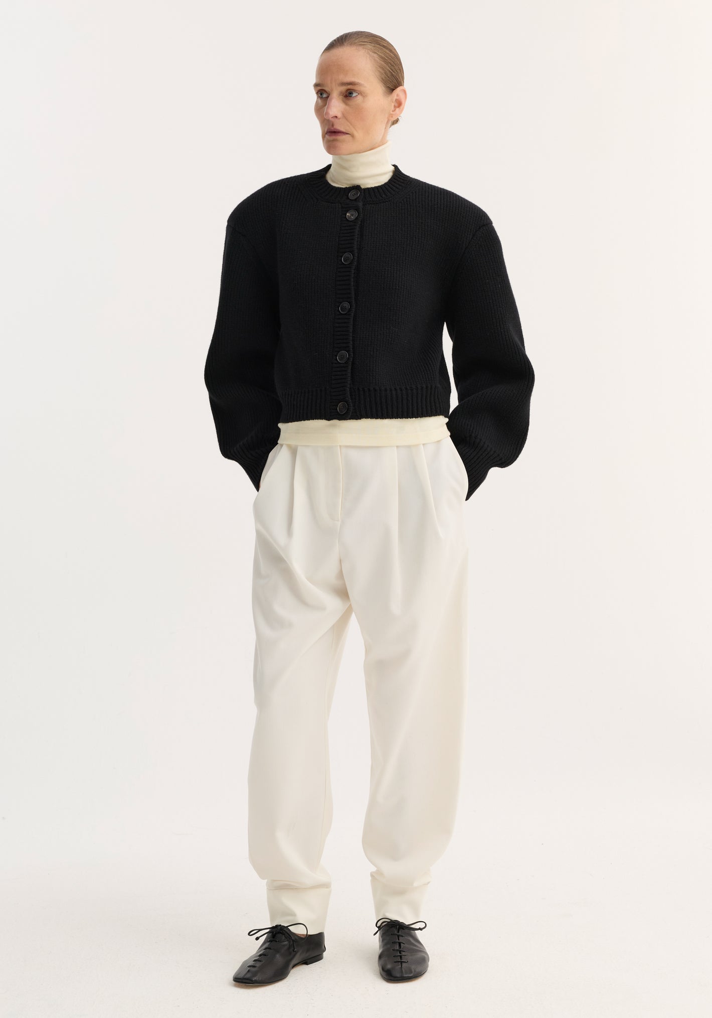 Model wearing Barrel leg trousers in off-white, featuring high-rise, relaxed tapered leg, double pleats, and ankle cuffs, styled with a black knit cardigan.