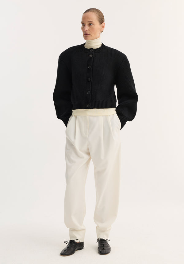 Model wearing Barrel leg trousers in off-white, featuring high-rise, relaxed tapered leg, double pleats, and ankle cuffs, styled with a black knit cardigan.